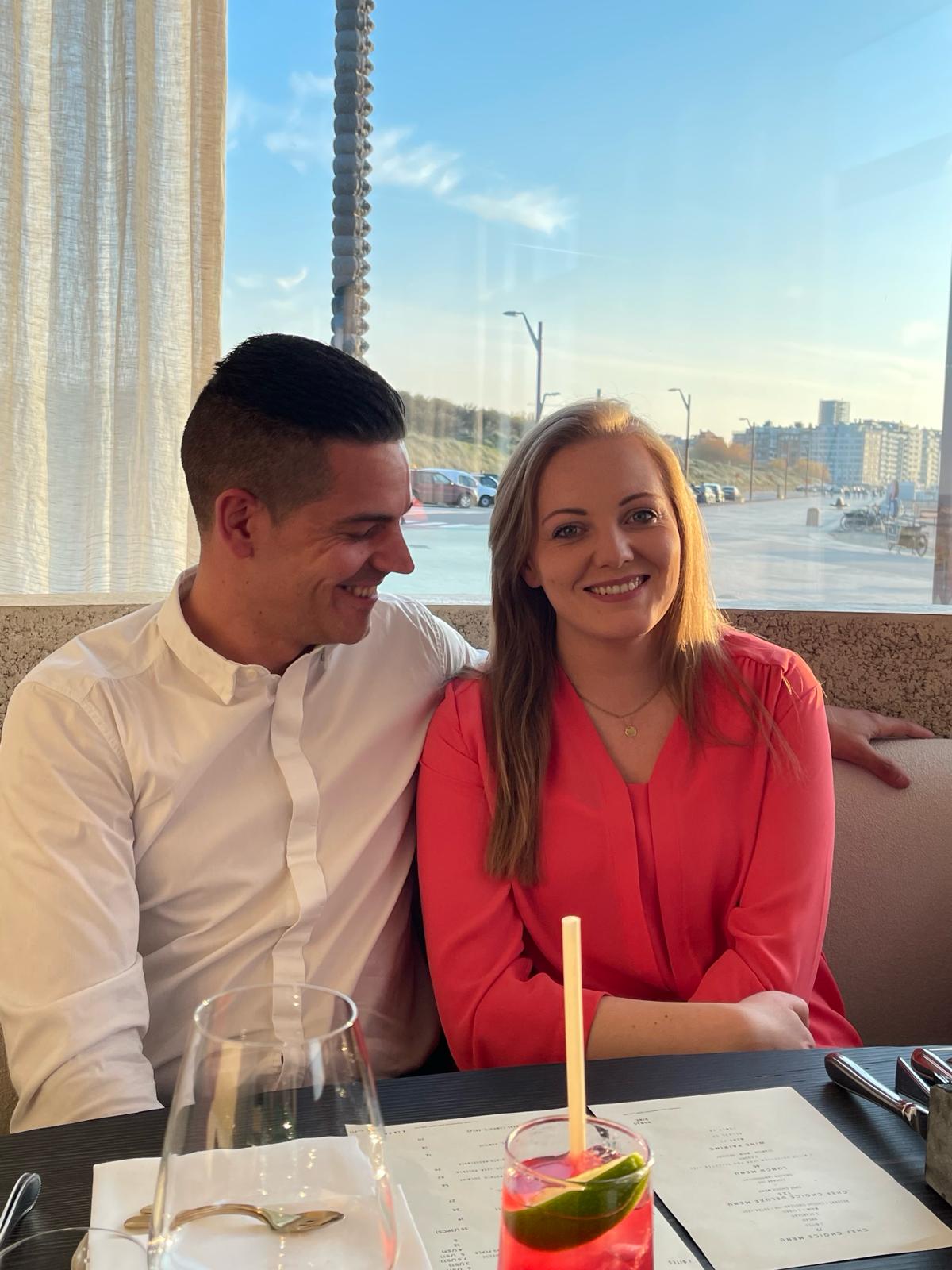Een koppel zit samen aan een restauranttafel bij een raam met uitzicht op een zonnige boulevard, met drankjes en menukaarten op tafel.