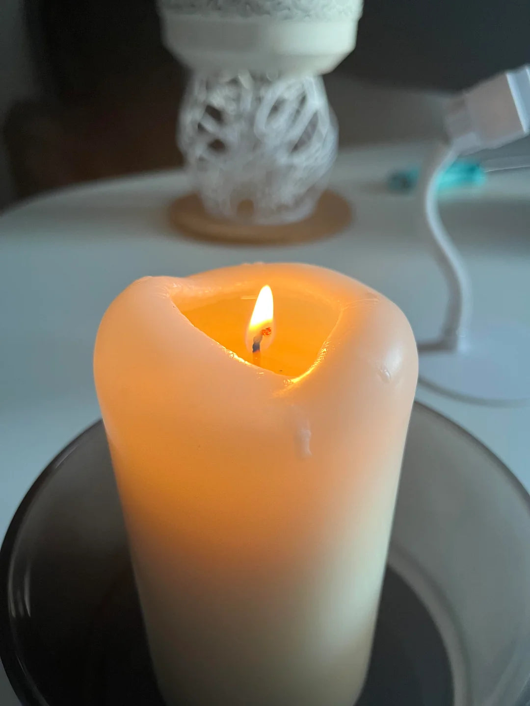 Brandende stompkaars met een ingezonken bovenkant, geplaatst in een donkere kaarshouder, op de achtergrond een decoratieve lamp en een oplaadkabel.