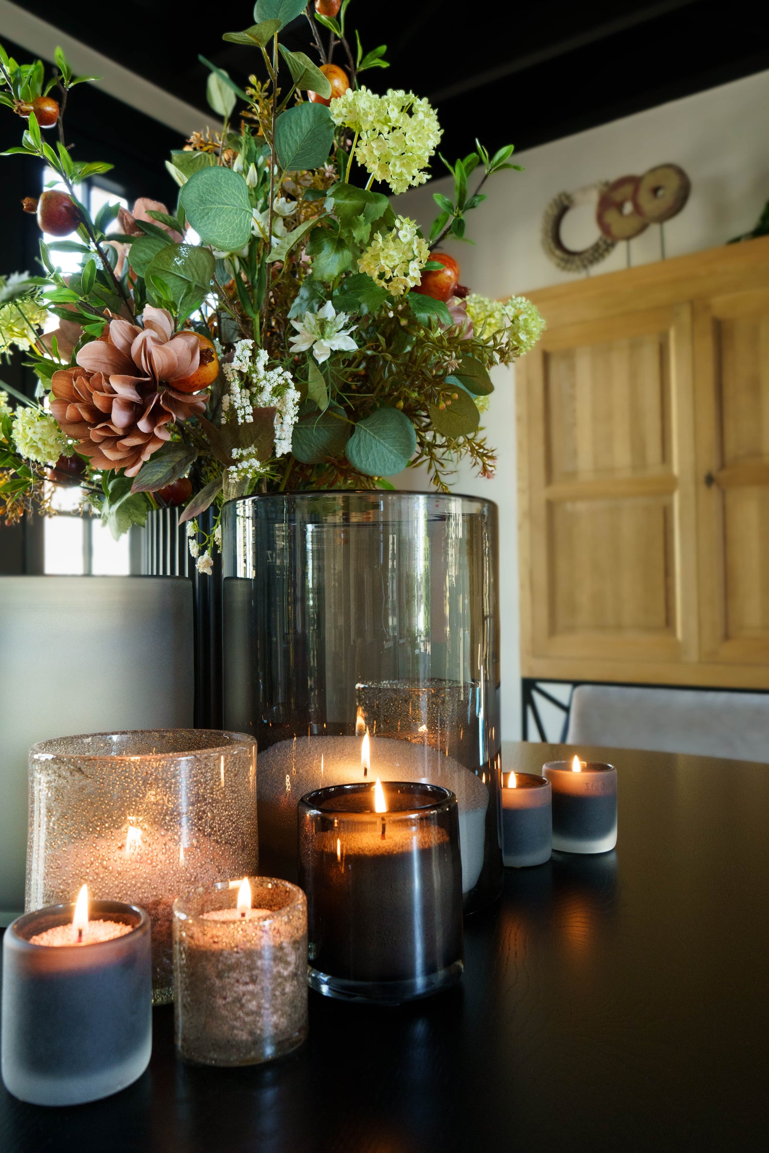 Een stijlvolle tafeldecoratie met meerdere brandende kaarsen in glazen houders in verschillende formaten en kleuren. Op de achtergrond staat een grote vaas met een bloemstuk van groene bladeren en gedroogde bloemen, wat een warme en natuurlijke sfeer creëert.