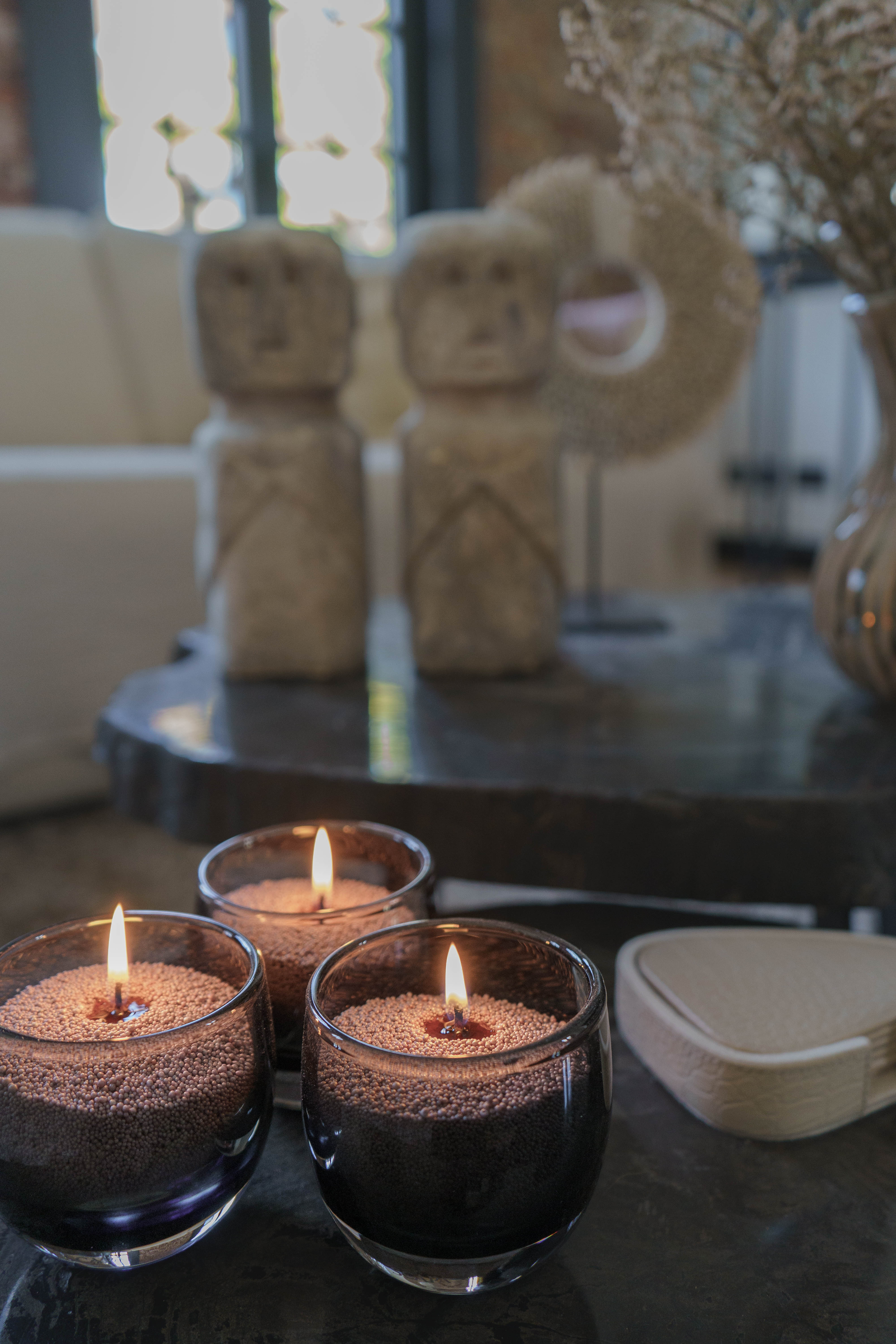 Drie brandende glazen kaarsen gevuld met bruin kaarsenzand staan op een donkere tafel. Op de achtergrond zijn decoratieve stenen beelden en een vaas met droogbloemen te zien, wat een warme en stijlvolle sfeer creëert.