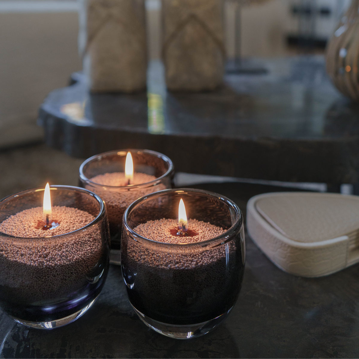 Drie brandende glazen kaarsen gevuld met bruin kaarsenzand van M1 Candles op een donkere tafel. De zachte gloed zorgt voor een warme, natuurlijke sfeer in het interieur.
