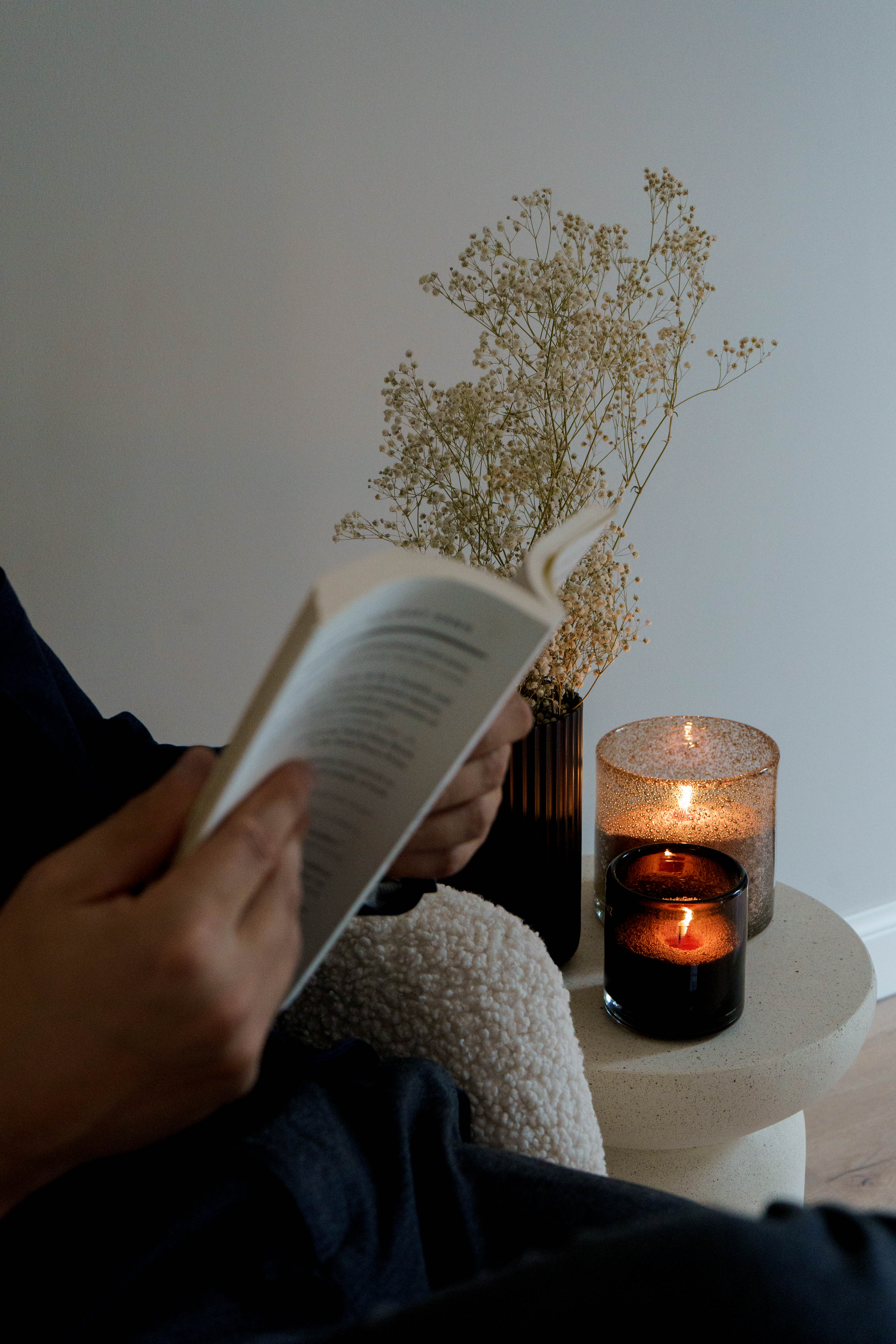 Een persoon leest ontspannen een boek naast twee brandende kaarsen in glazen houders. Op de achtergrond staat een vaasje met gedroogde bloemen, wat een warme en rustige sfeer creëert.