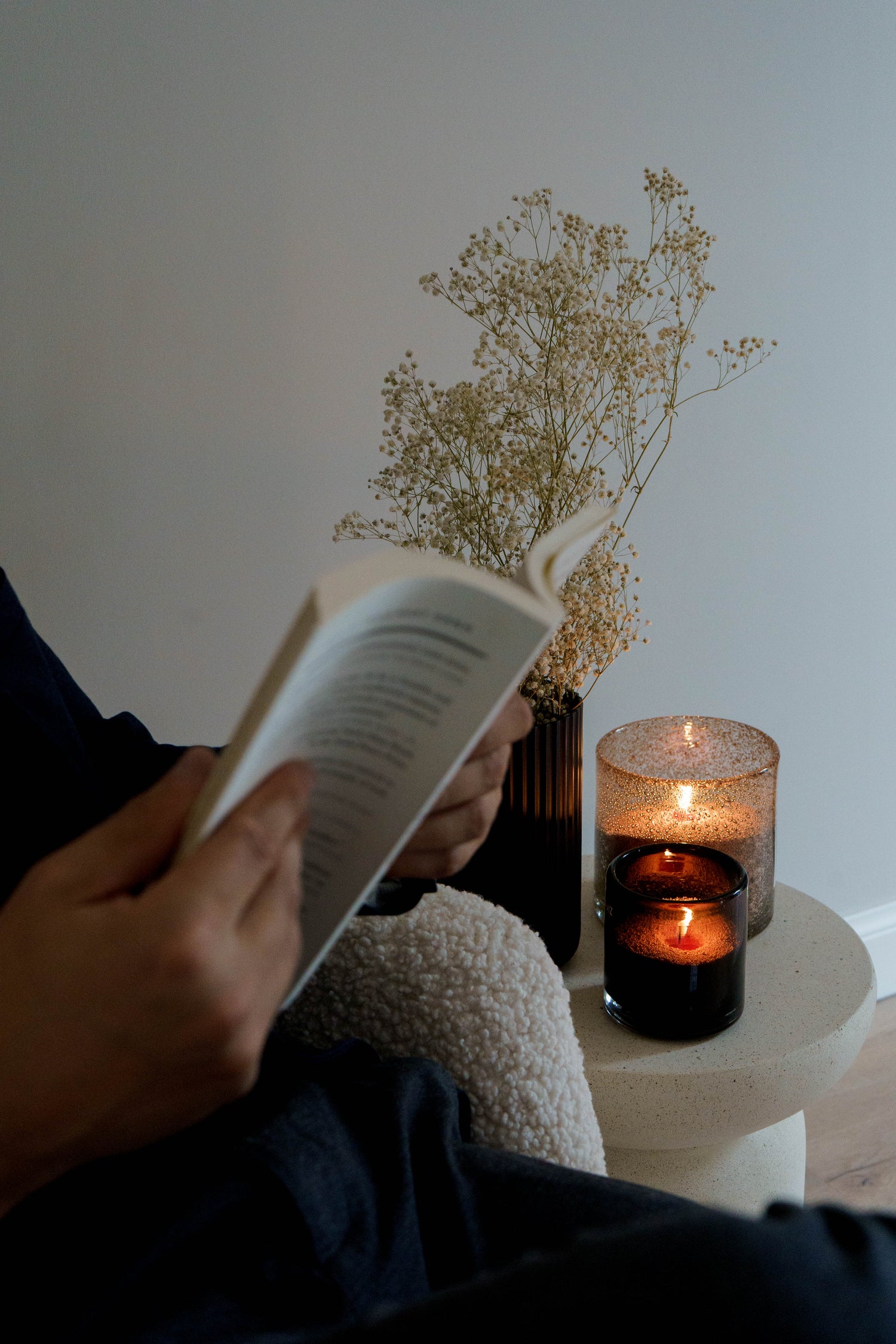 Een persoon leest ontspannen een boek naast twee brandende kaarsen in glazen houders. Op de achtergrond staat een vaasje met gedroogde bloemen, wat een warme en rustige sfeer creëert.
