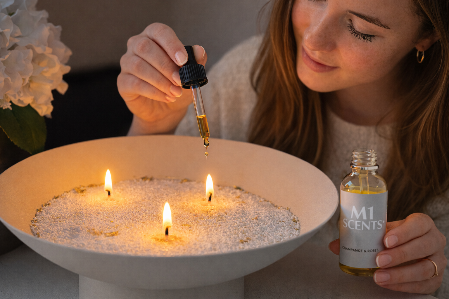 Close-up van kaarsenzand met drie lonten terwijl een vrouw M1 Scents geurolie toevoegt voor een intensere geur en sfeervolle verlichting.