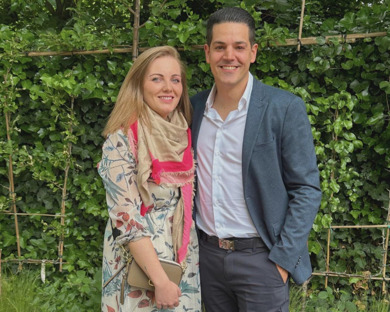 Een koppel poseert buiten voor een groene haag; de vrouw draagt een bloemenjurk met een sjaal en de man een donkerblauw colbert met witte blouse.