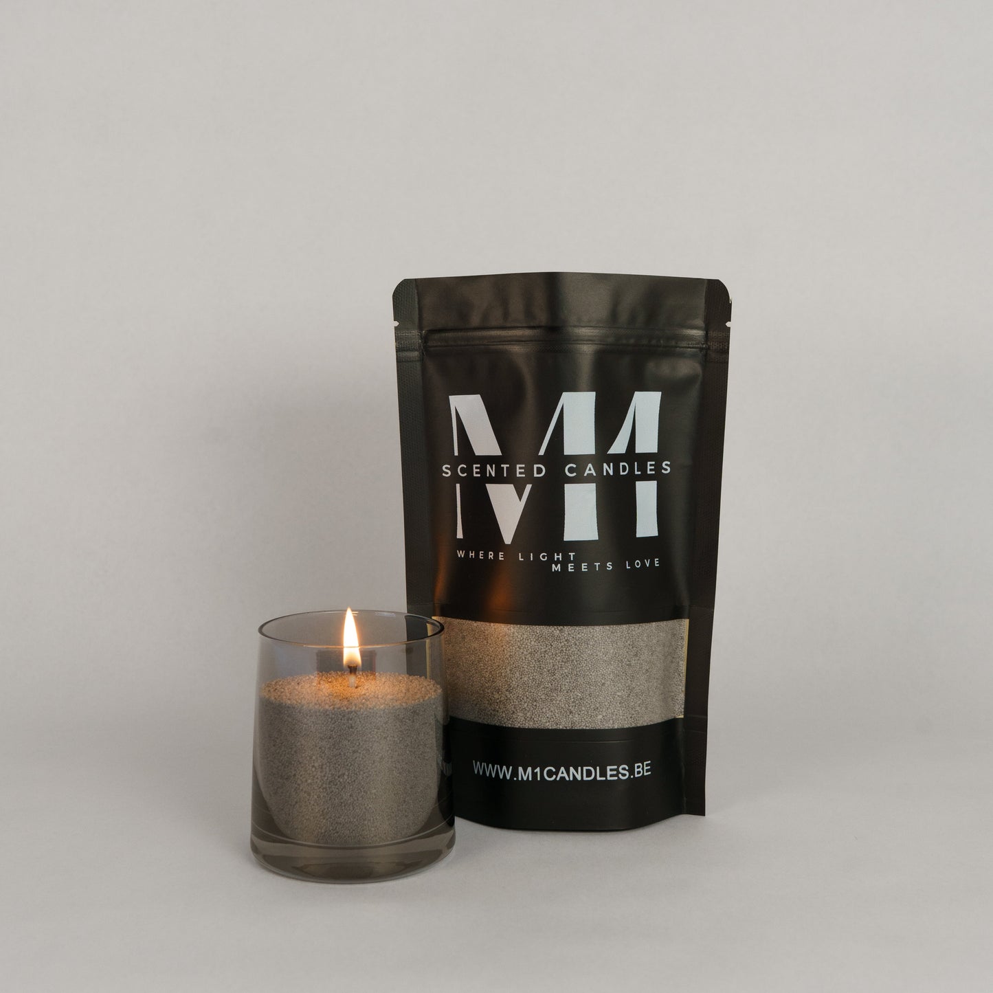 Verpakking van M1 Scented Candles kaarsenzand in een zwarte zak met logo, naast een glazen houder gevuld met brandend grijs kaarsenzand.