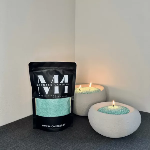 M1 Candles verpakking met turquoise kaarsenzand, sfeervol opgesteld naast twee brandende kaarsen in decoratieve stenen kommen, die samen een gezellige en ontspannen sfeer creëren.